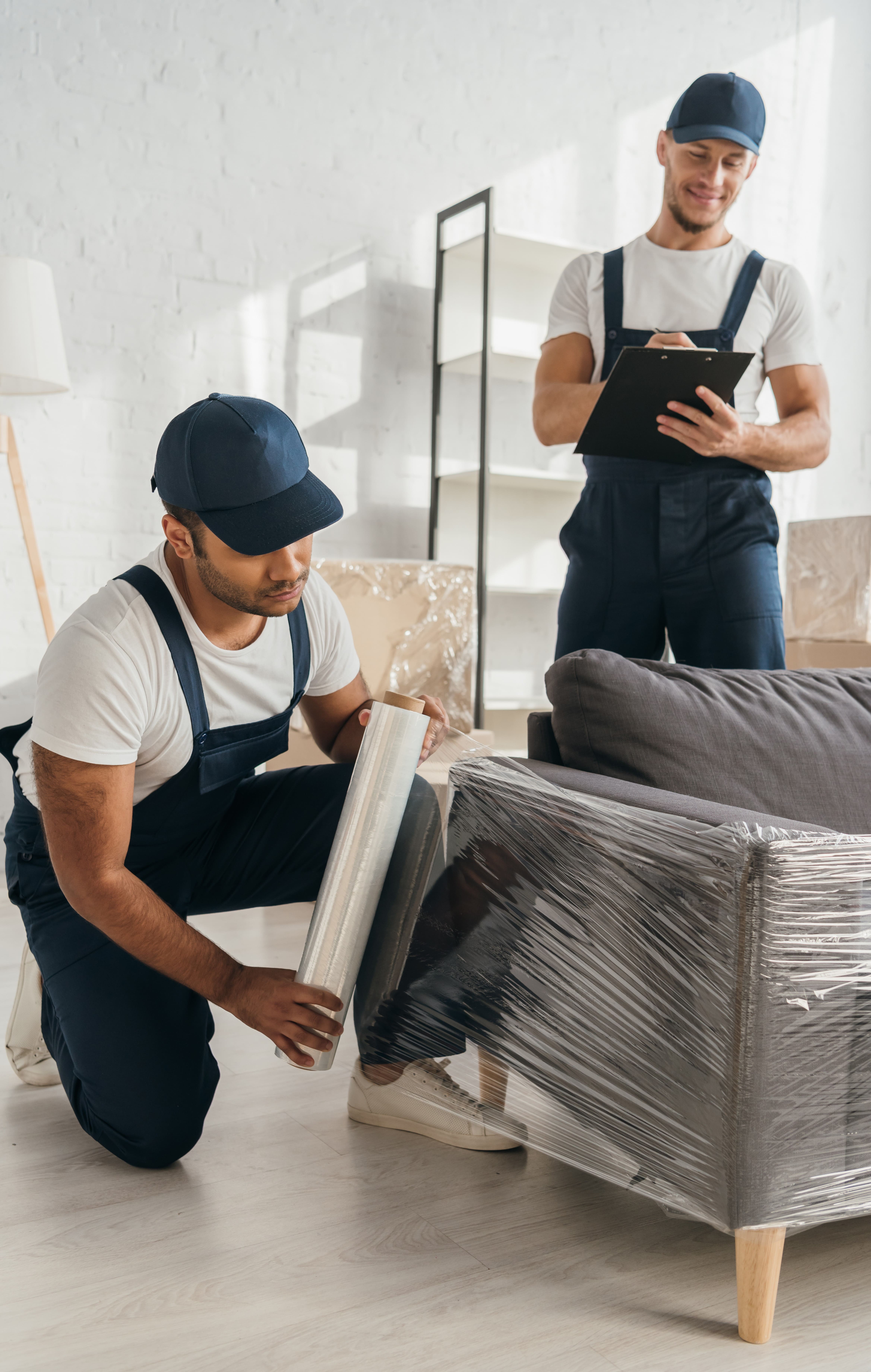 Indian mover holding stretch film roll near sofa with a worker checking details on a clipboard in the background