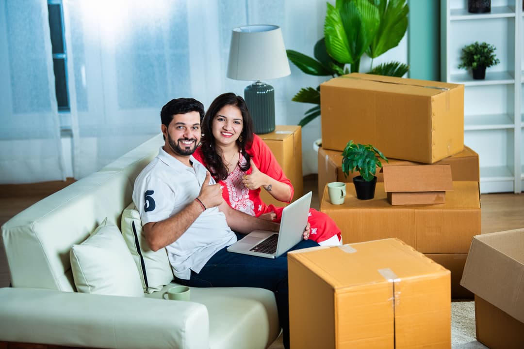 Young Indian couple using a laptop on a sofa amidst home shifting with moving boxes around